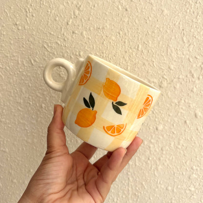 A hand holding a white ceramic mug with a yellow-orange lemon and orange pattern against a textured beige wall.