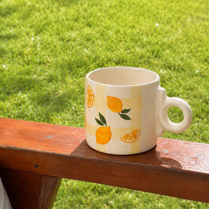 A cream-colored ceramic mug with a yellow and white gingham pattern and a lemon print, resting on a wooden armchair outdoors on a green grass lawn.