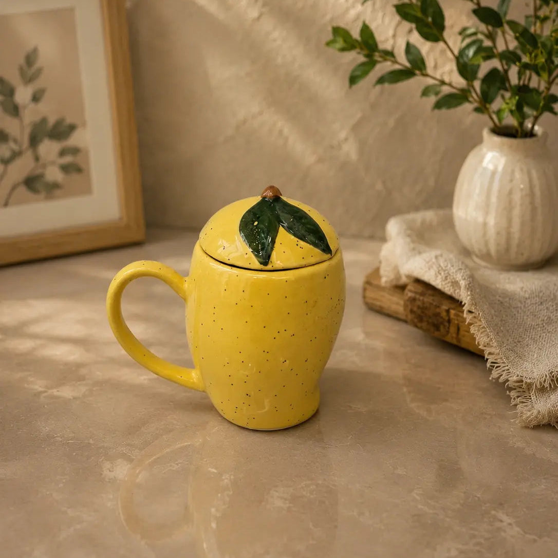 Yellow ceramic mango shaped mug with a lid on a textured surface with a plant and framed picture in the background.