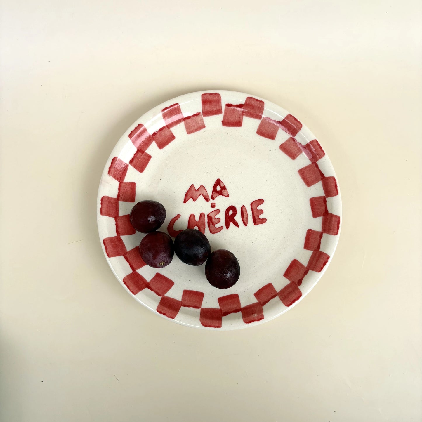 A white ceramic plate with a red checkered border and the words 'MA CHÉRIE' written in red on the center, holding three dark red cherries, on a plain beige background.