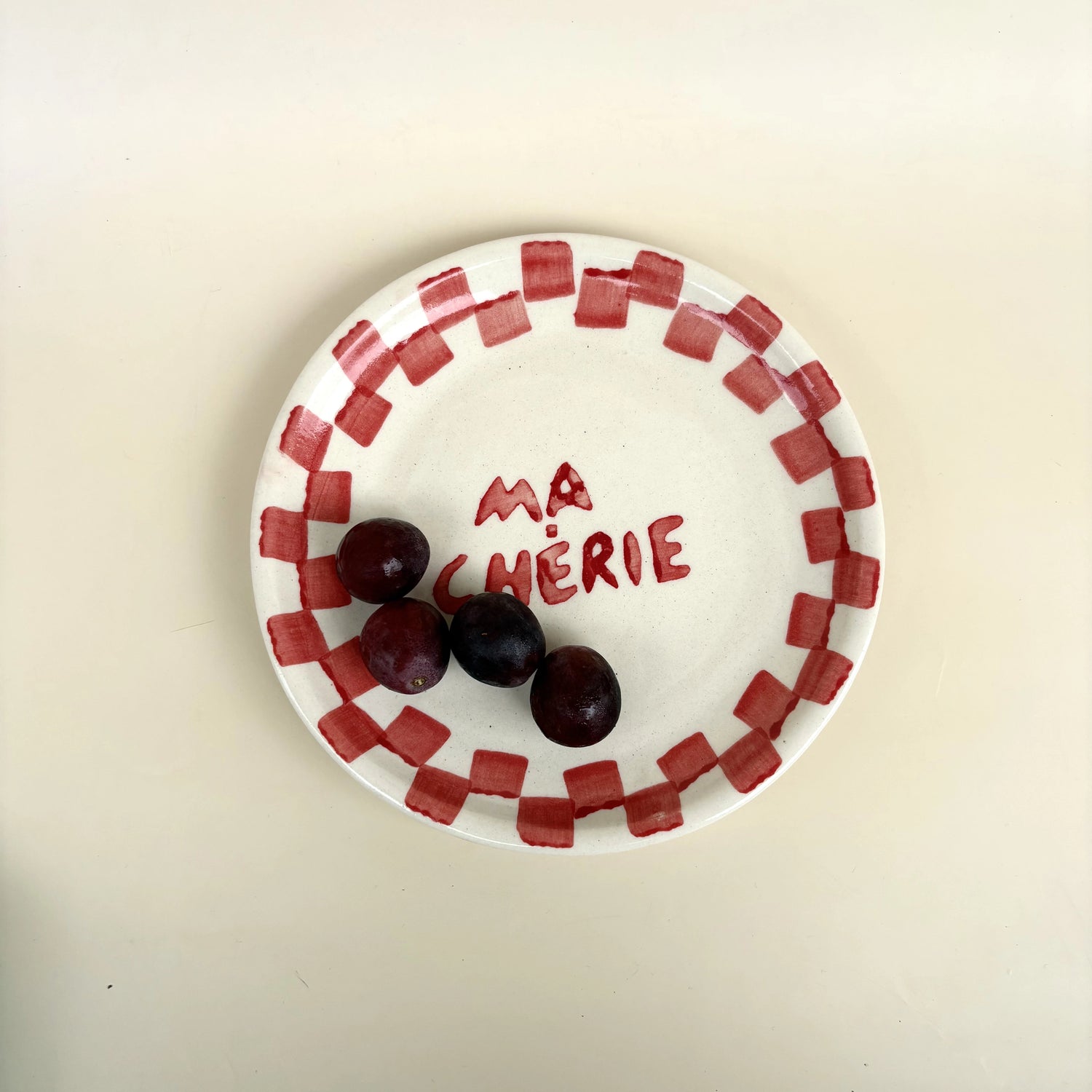 A white ceramic plate with a red checkered border and the words 'MA CHÉRIE' written in red on the center, holding three dark red cherries, on a plain beige background.