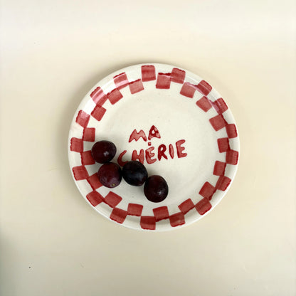 A white ceramic plate with a red checkered border and the words 'MA CHÉRIE' written in red on the center, holding three dark red cherries, on a plain beige background.