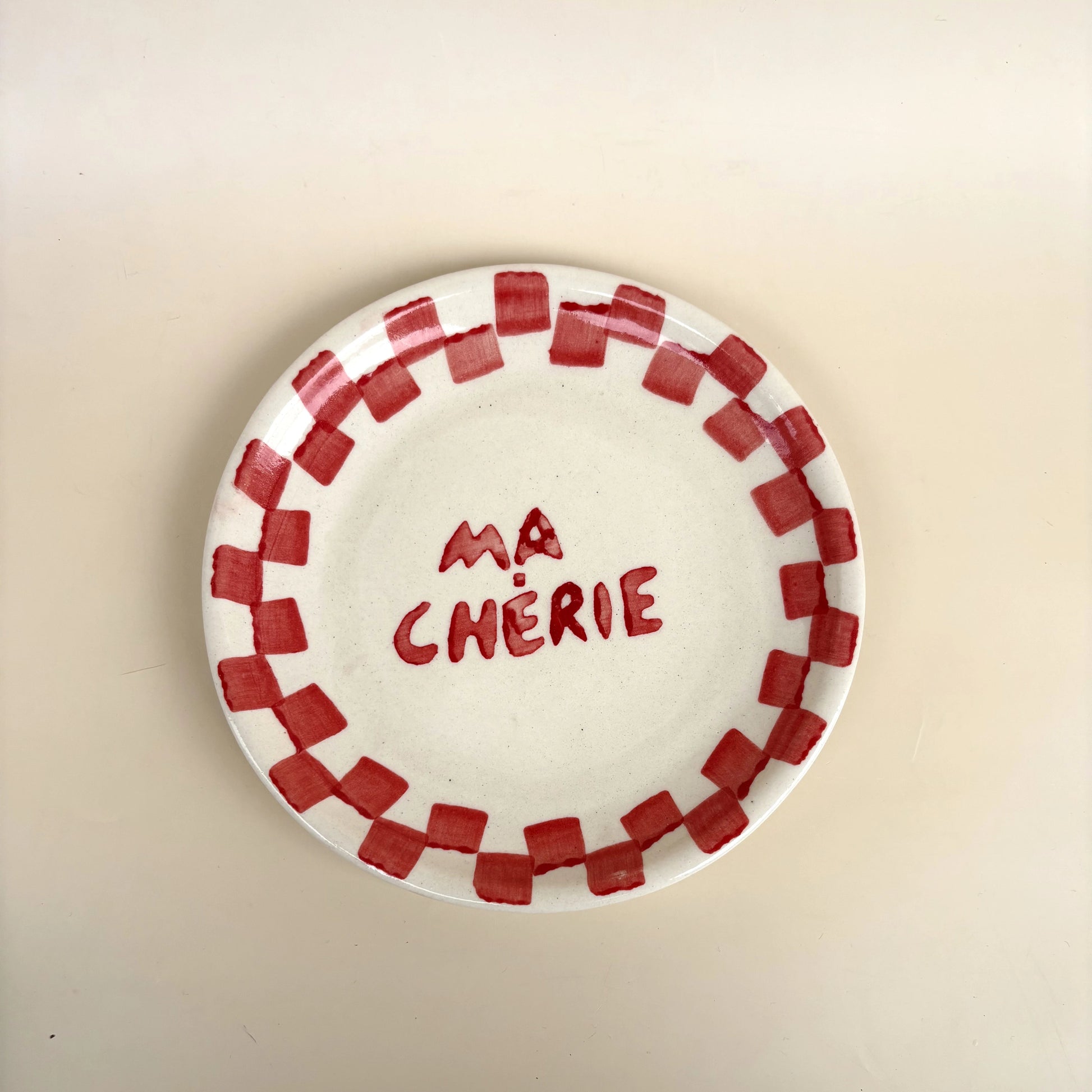 A white ceramic plate with a red checkered border and the French text 'MA CHÉRIE' written in red in the center, shown on a plain beige background.