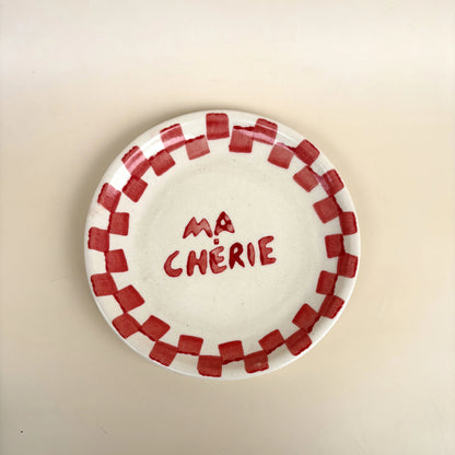 A white ceramic plate with a red checkered border and the French text 'MA CHÉRIE' written in red in the center, shown on a plain beige background.