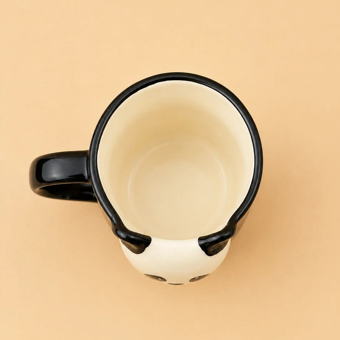 A top-down view of a ceramic mug designed to look like a panda face, featuring a black handle, black rim, and black ears on a cream-colored body, set against a solid beige background.