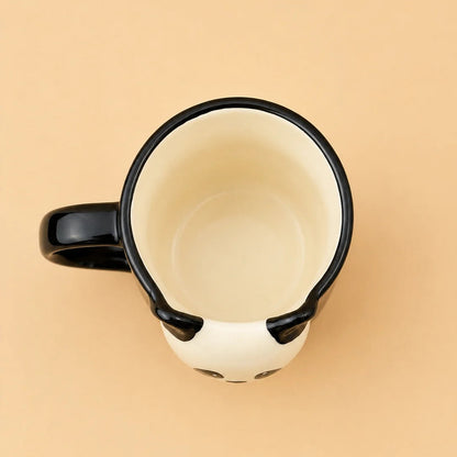 A top-down view of a ceramic mug designed to look like a panda face, featuring a black handle, black rim, and black ears on a cream-colored body, set against a solid beige background.