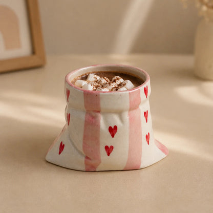A small ceramic mug shaped like a fabric pouch, decorated with pink vertical stripes and red hearts, filled with a hot beverage topped with marshmallows, set on a beige surface with soft background shadows.