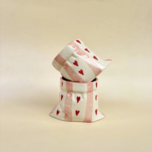 A small decorative ceramic container with a matching lid, featuring a white and light pink striped pattern with small red hearts, set against a plain beige background.