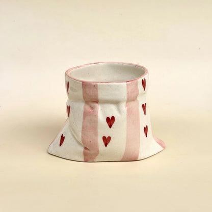 A handmade ceramic cup with a flared base, decorated with vertical pink stripes and small red hearts on a cream-colored glaze, set against a solid cream background.
