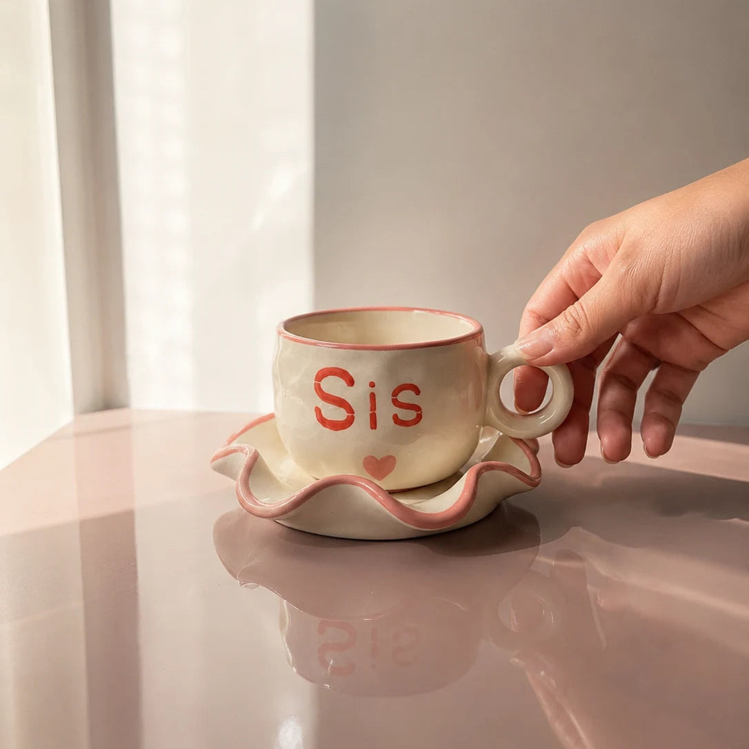 A hand holds a beige ceramic teacup and saucer set with a pink rim and the word 'Sis' in red on the cup, set on a glossy pink surface against a soft neutral background.