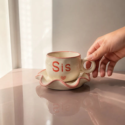 A hand holds a beige ceramic teacup and saucer set with a pink rim and the word 'Sis' in red on the cup, set on a glossy pink surface against a soft neutral background.