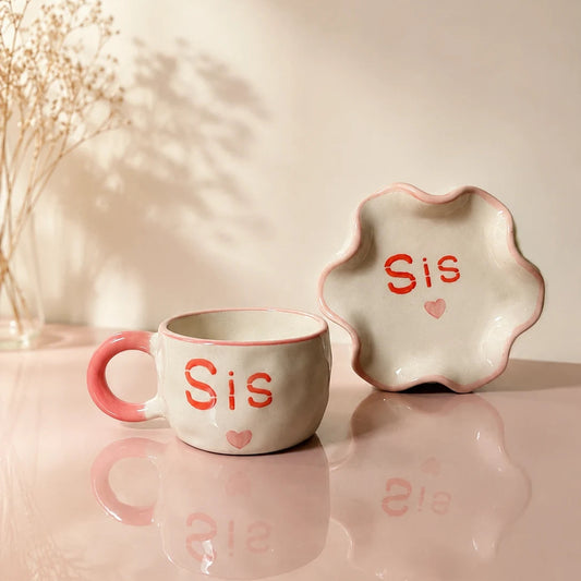 A ceramic mug and matching flower-shaped plate in a soft cream color with coral-pink lettering that reads 'Sis' and a small heart, styled on a reflective pink surface with dried flowers in the background.