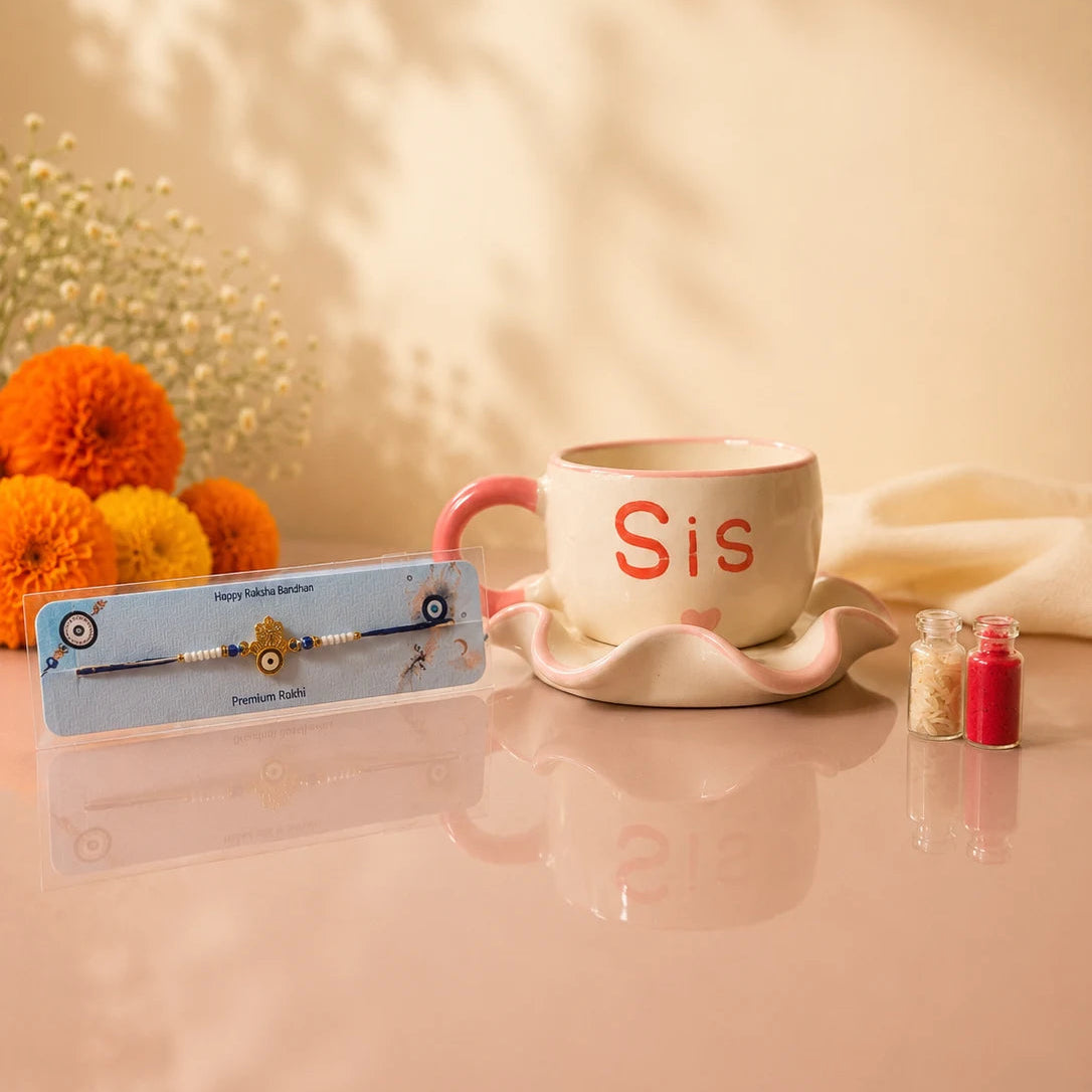 A framed traditional Indian crystal and bead bracelet with a gold-toned 'Sis' nameplate is displayed on a reflective pink surface next to a pink and white ceramic mug with the word 'Sis' in red, decorated with orange marigold flowers and soft shadow patterns on a warm beige background.