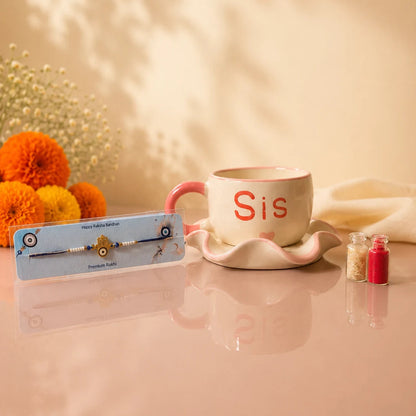 A framed traditional Indian crystal and bead bracelet with a gold-toned 'Sis' nameplate is displayed on a reflective pink surface next to a pink and white ceramic mug with the word 'Sis' in red, decorated with orange marigold flowers and soft shadow patterns on a warm beige background.