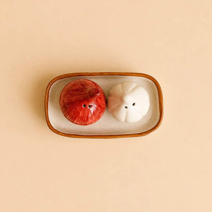 Two ceramic salt and pepper shakers shaped like a red onion and a garlic bulb, resting in a small rectangular ceramic dish on a solid beige background.