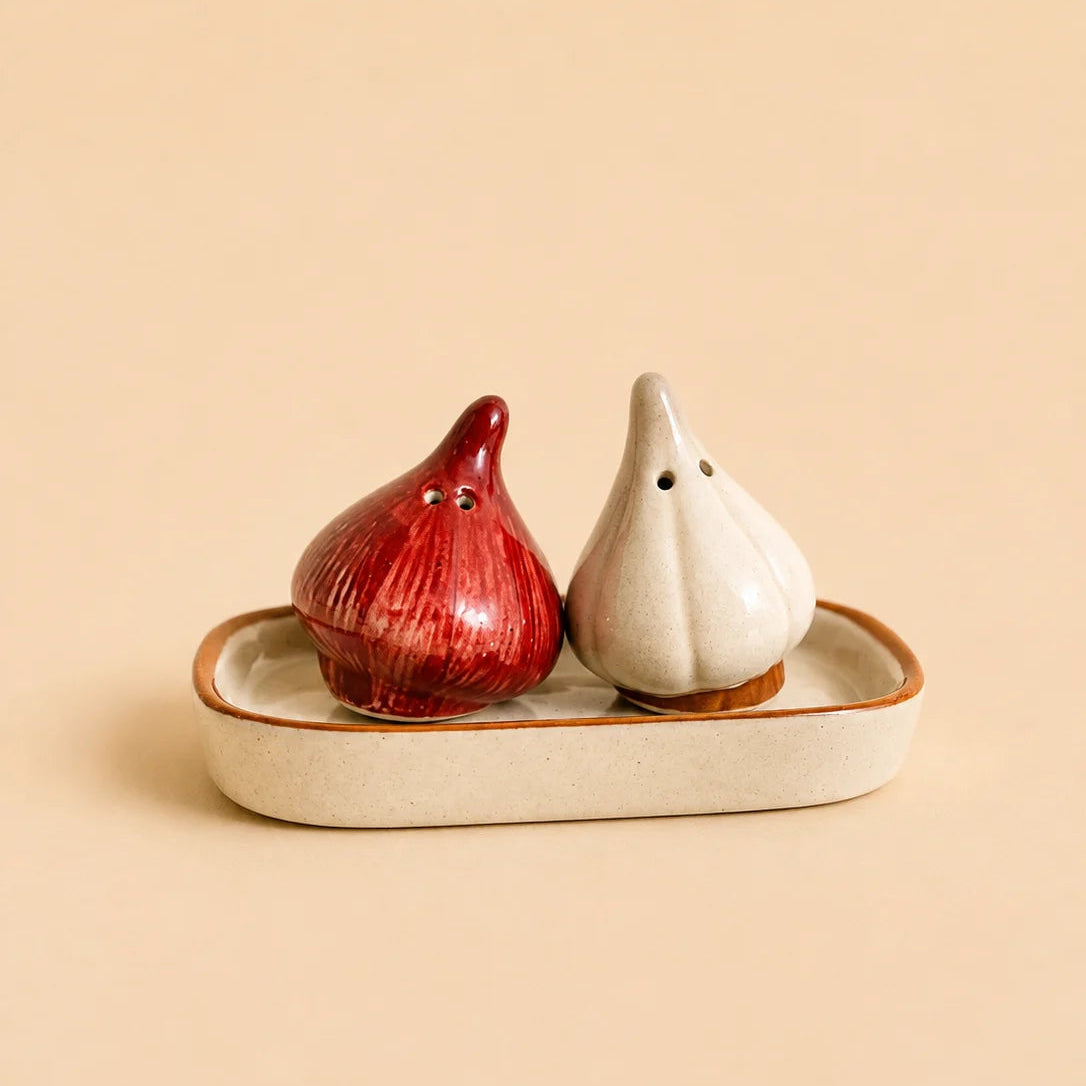 Two ceramic garlic-shaped salt and pepper shakers, one red and one off-white, sitting on a matching beige ceramic tray against a solid beige background.