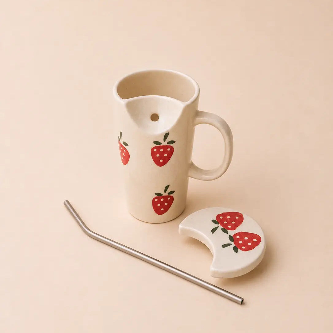 A white ceramic pitcher with a handle and spout, decorated with red strawberry illustrations, shown with a matching strawberry-patterned lid and a bent metal straw on a warm beige background.