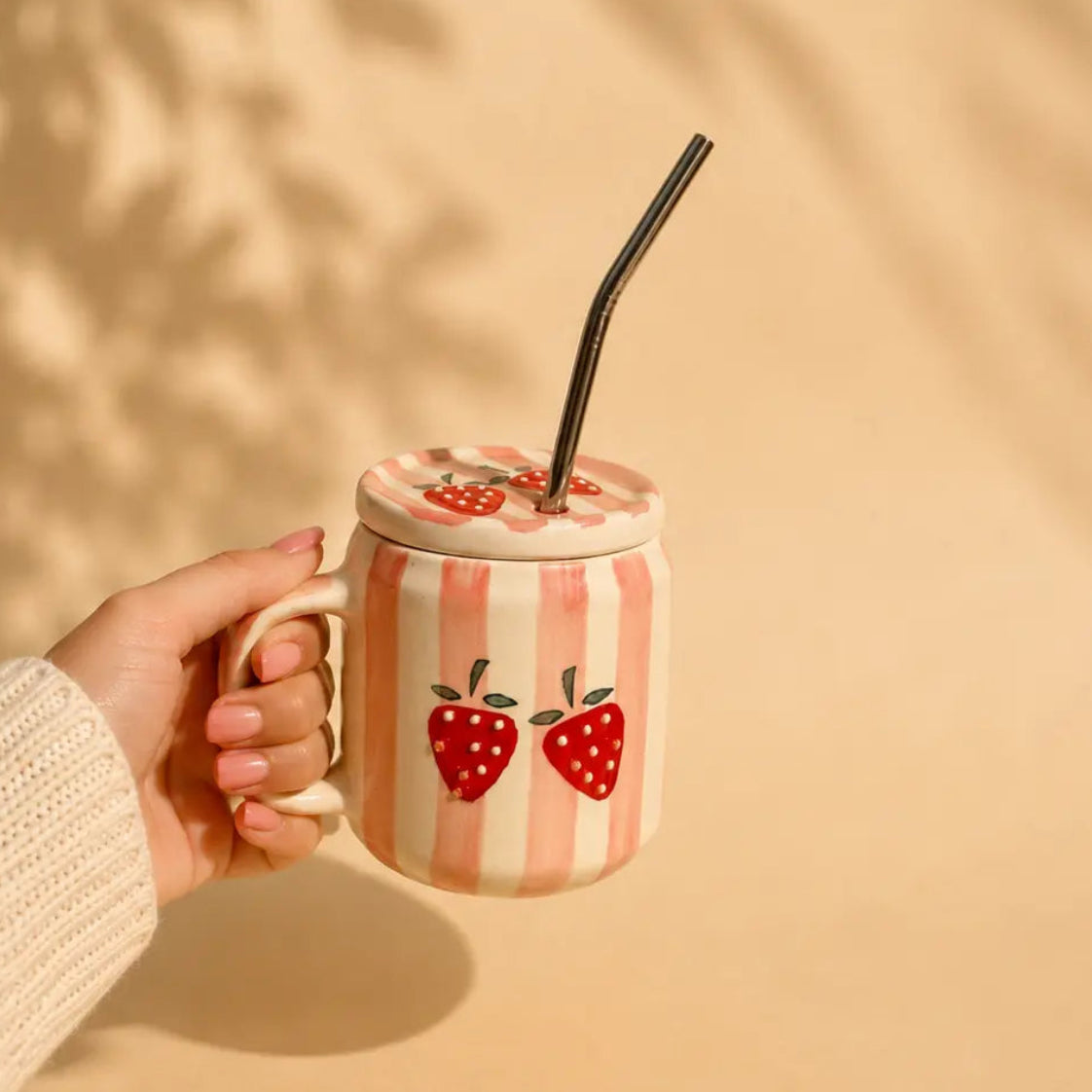 A hand in a white knit sleeve holds a pink and white striped ceramic mug with two red strawberry illustrations. The mug has a matching lid with a black straw, set against a warm beige background with soft leaf shadows.