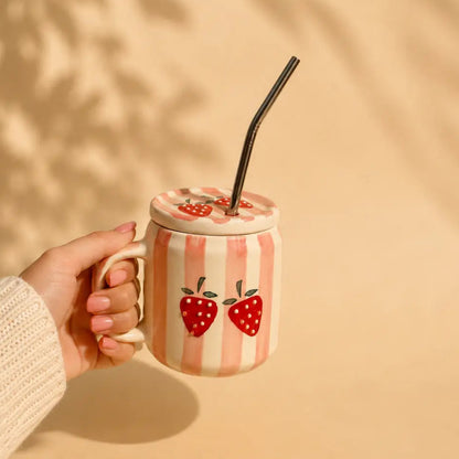 A hand in a white knit sleeve holds a pink and white striped ceramic mug with two red strawberry illustrations. The mug has a matching lid with a black straw, set against a warm beige background with soft leaf shadows.
