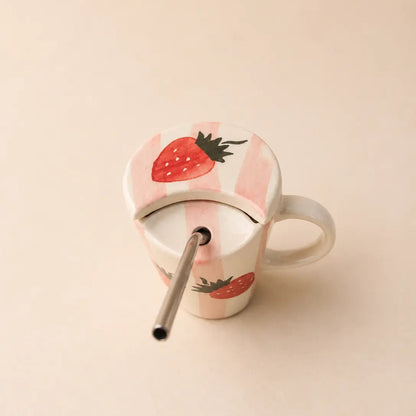 A ceramic mug with a matching lid and metal straw, decorated with a pink and white striped pattern and red strawberry illustrations, set against a solid beige background.