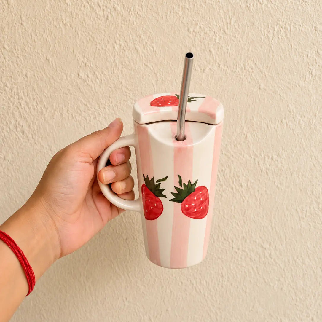 A hand holding a pink and white striped insulated sipper with a lid, stainless steel straw, and strawberry illustrations, set against a textured beige wall.