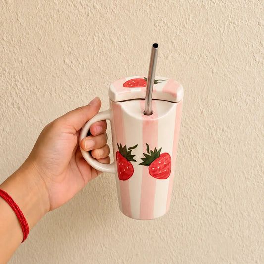 A hand holding a pink and white striped insulated sipper with a lid, stainless steel straw, and strawberry illustrations, set against a textured beige wall.