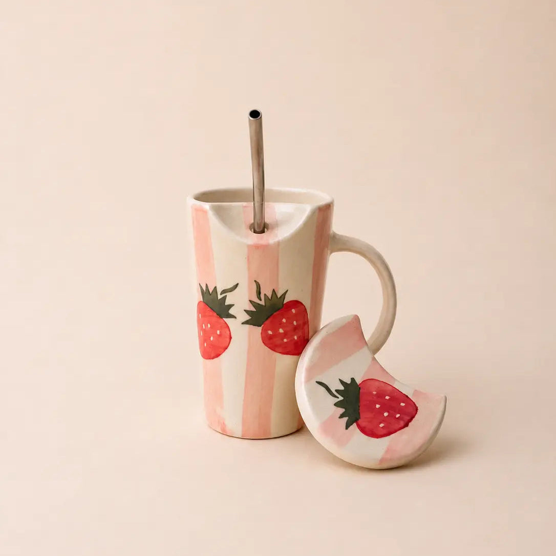 A pastel pink and white striped ceramic mug with painted red strawberries, shown with a matching strawberry-themed lid and a metal straw on a warm beige background.