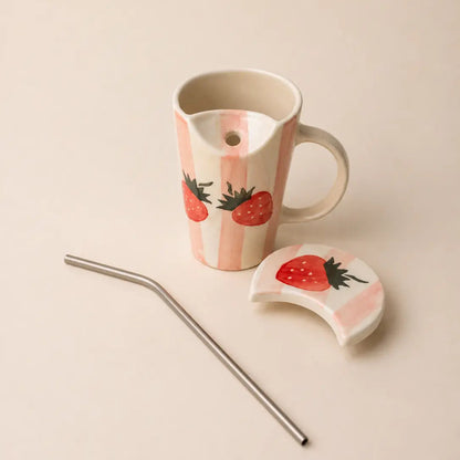 A ceramic mug with a handle, decorated with pink and white vertical stripes and red strawberries, shown with a matching lid and a metal straw on a light beige background.