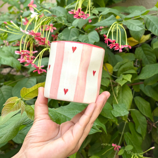 Hand holding a small ceramic pot with red heart patterns against a green leafy background